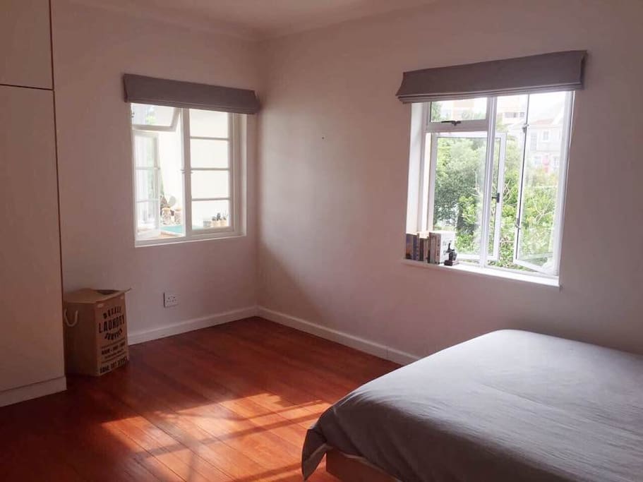 The bedroom: view of the sunny space with two windows, queen-sized bed, laundry basket and cupboard space.