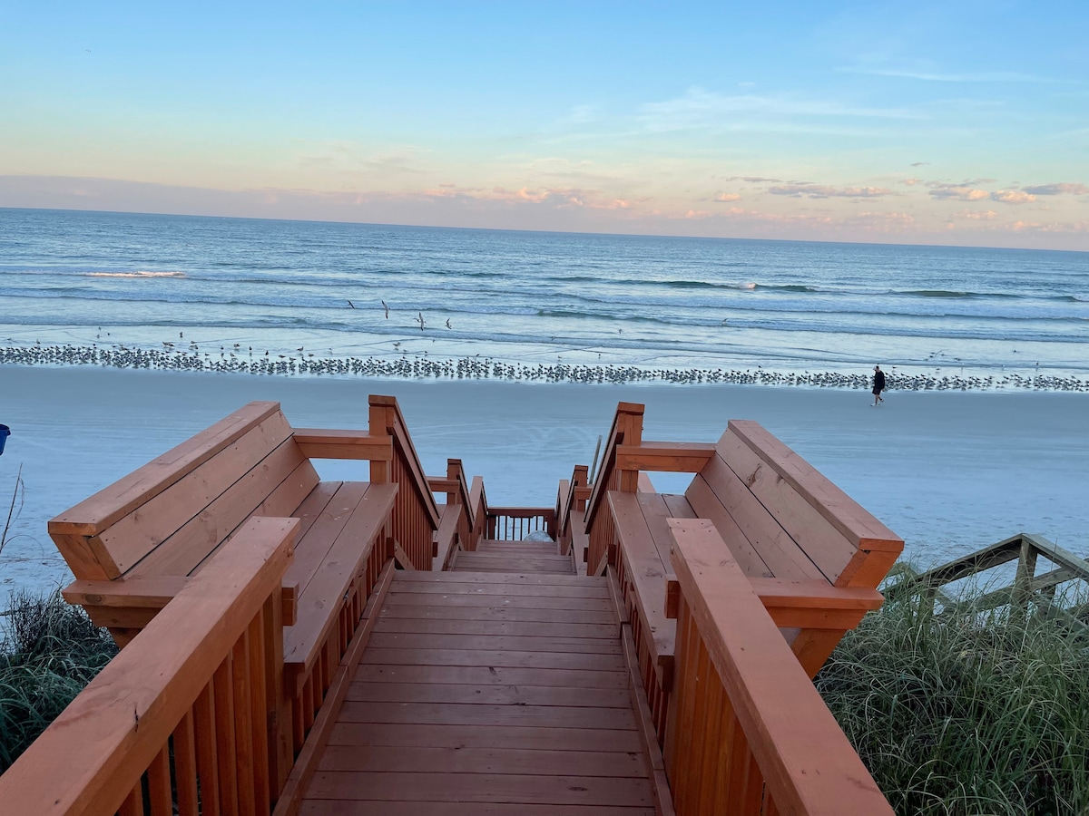 A wooden staircase descends toward the sandy beach, constructed with broad steps and railings. The ocean is visible in the background, with gentle waves and a clear sky. Shorebirds can be seen along the water's edge, complementing the tranquil coastal scene.