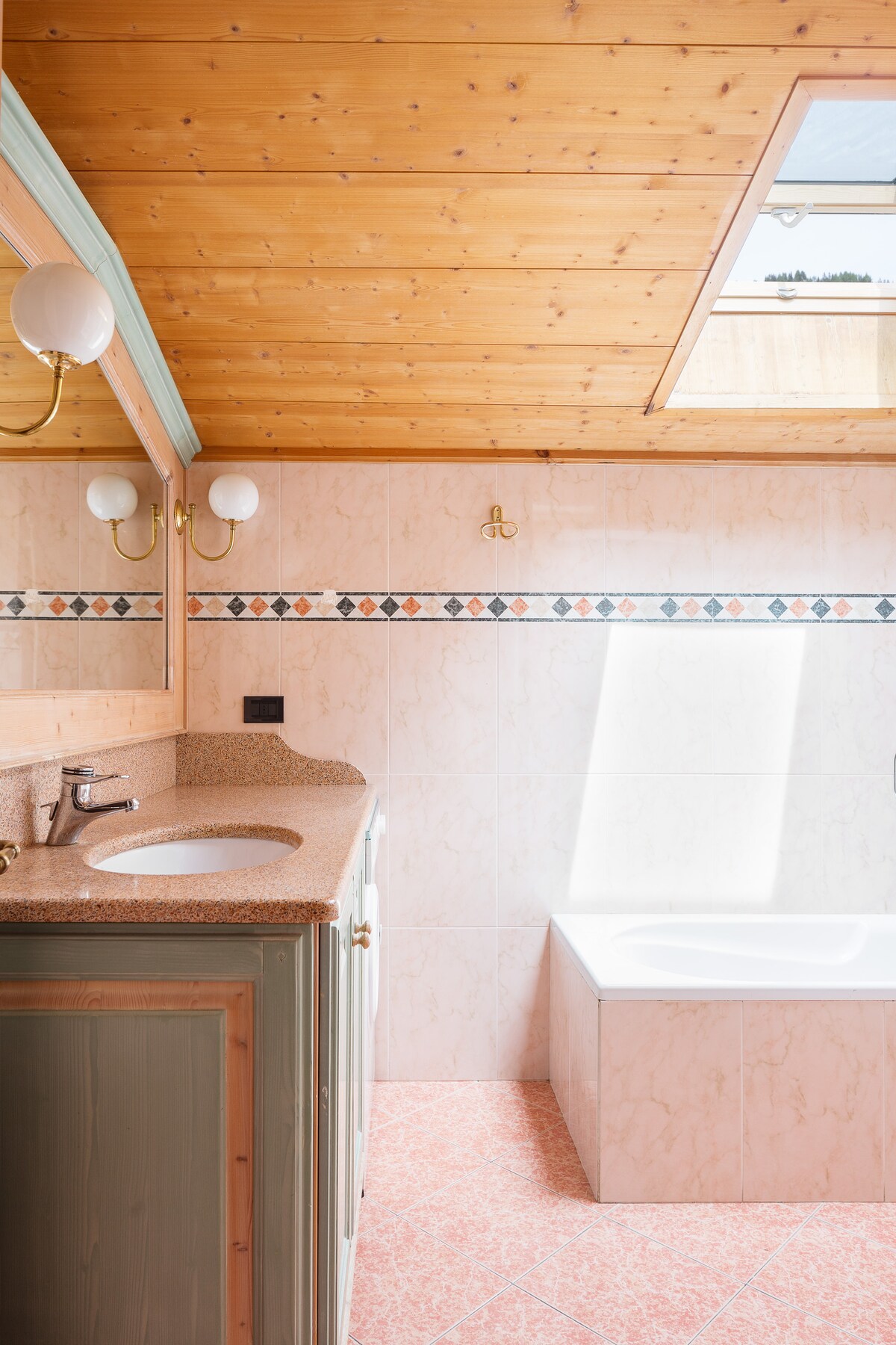 A bathroom features a light-colored tiled wall and a bathtub with a combination of a wooden ceiling and a skylight above. The countertop is finished in a neutral tone, complemented by brass fixtures and enclosed lighting, creating a bright and airy space.