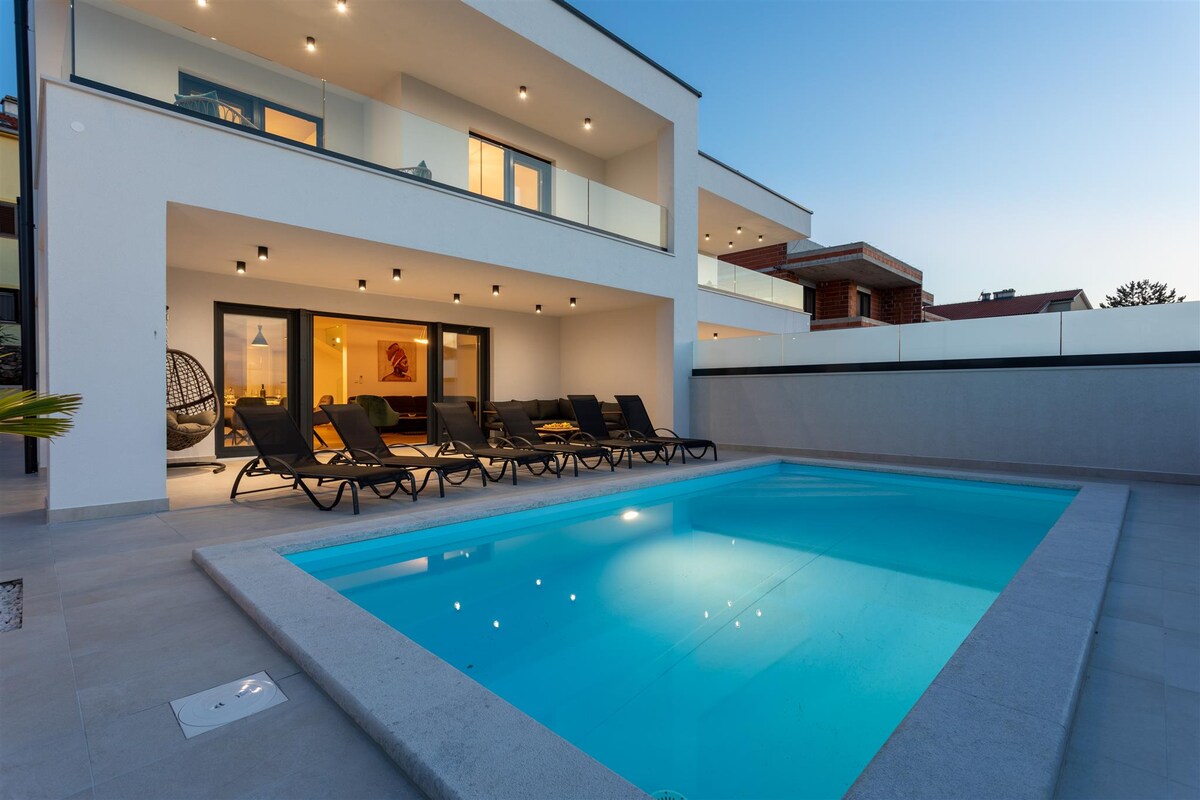 The outdoor area showcases a heated pool surrounded by eight sun loungers, all positioned for relaxation. The modern villa features large glass doors that provide views into the brightly lit interior. Soft lighting enhances the tranquil atmosphere as evening falls.