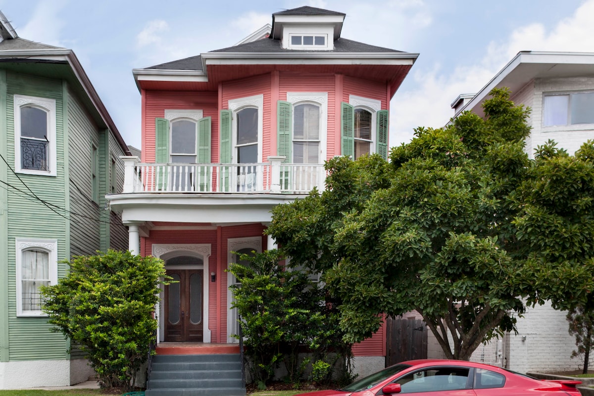 A Victorian-style building with a vibrant red and white facade features green shutters on its bay windows. A welcoming porch with white railing extends along the front. The well-maintained garden is lush with green foliage, contributing to the charm of the entrance.