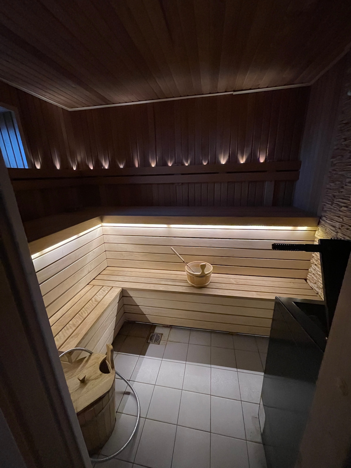 A warm wooden sauna is featured, with smooth benches lining the walls and soft lighting creating a calming effect. A wooden bucket sits nearby, with a metal stove visible in the corner. The tiled floor contrasts with the warm wood tones.