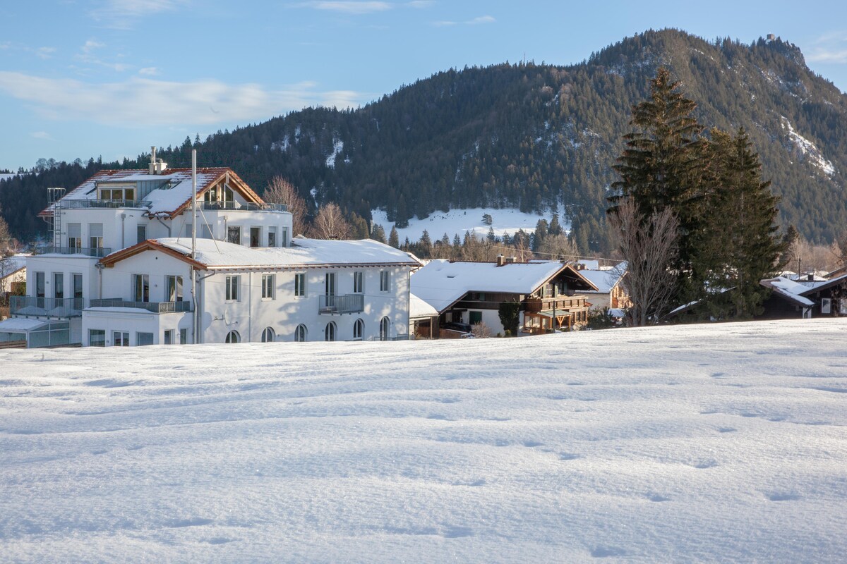 The exterior of a three-story building is shown, with white walls contrasting against a snowy landscape. Surrounding mountains are visible in the background, and nearby wooden chalets add to the serene winter scene. The ground is covered in fresh snow, reflecting sunlight.