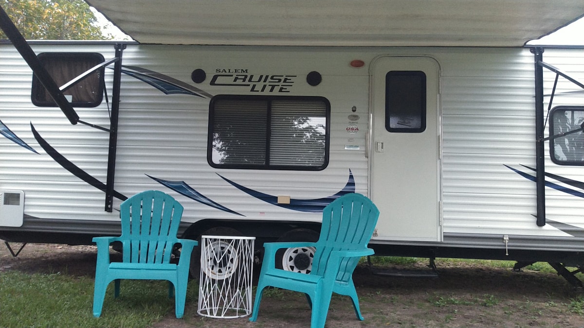 A compact exterior of a travel trailer is shown, featuring a light-colored shell with decorative blue accents. Two turquoise chairs are positioned near a small white table, inviting relaxation in the outdoors. An awning extends from the trailer, providing shade.