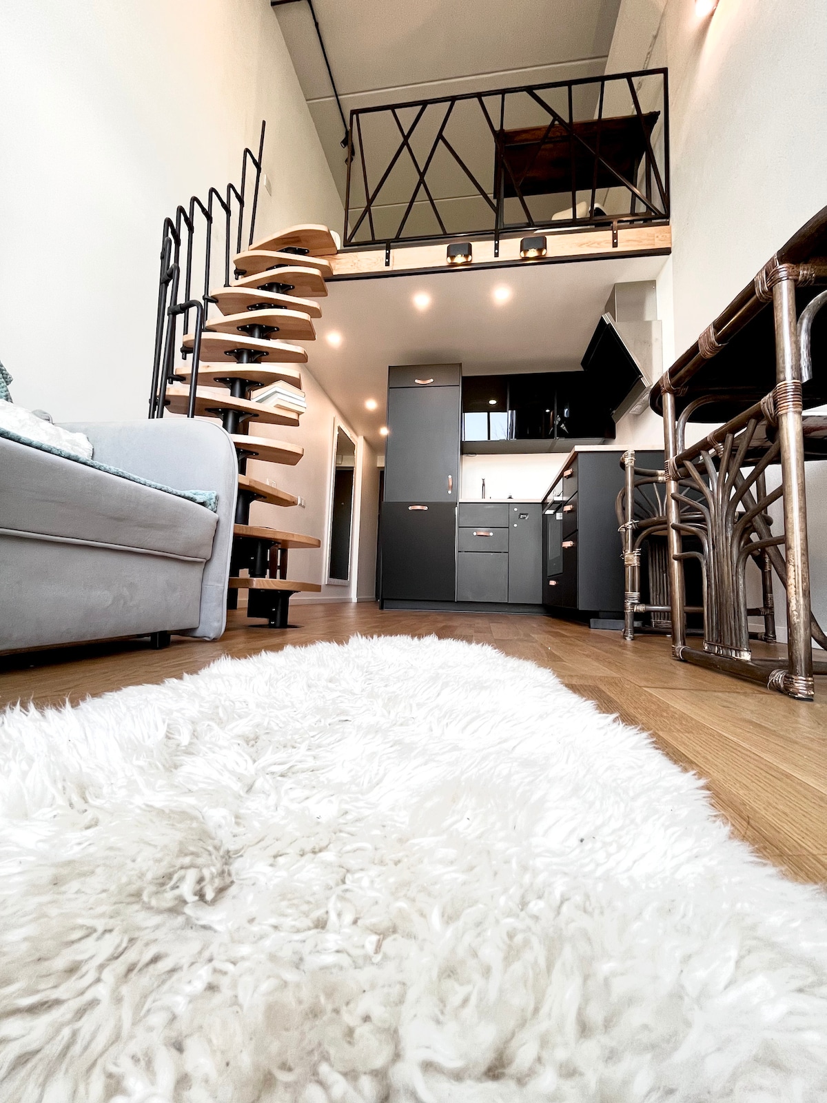The image captures a modern loft interior featuring a spiral staircase leading to a lofted area. A cozy rug is positioned on wooden flooring, enhancing the inviting layout. A compact kitchen is visible, characterized by dark cabinetry. Natural light illuminates the space.