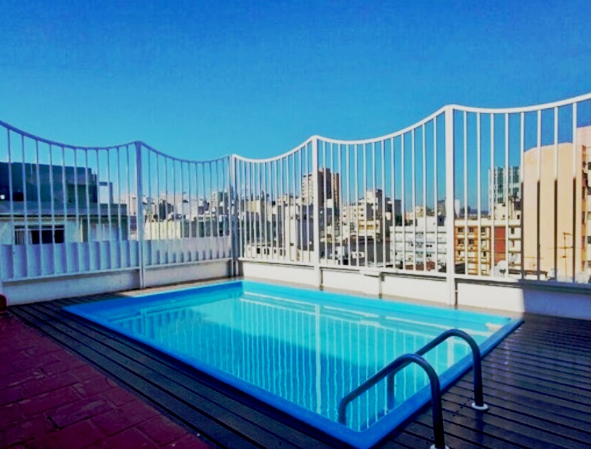 A rooftop pool is surrounded by a white railing, offering views of the city skyline. The pool is rectangular, with a light blue surface reflecting the clear sky above. A wooden deck frames the pool area, enhancing the outdoor space.