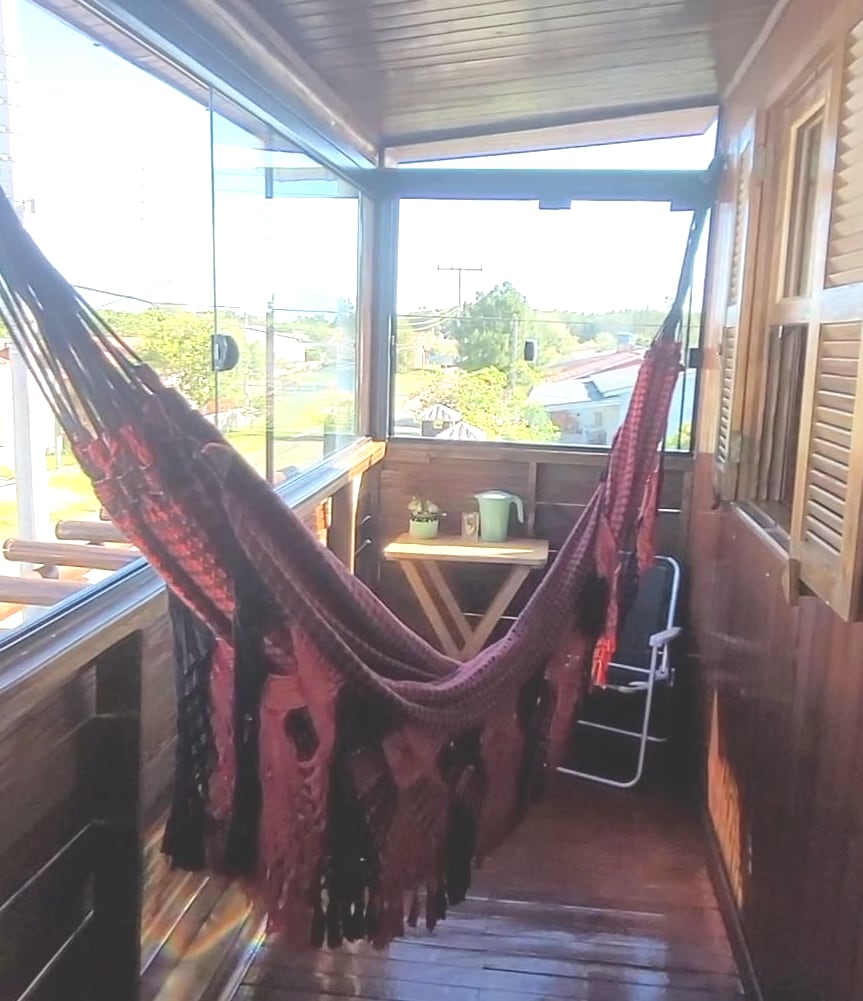 A cozy balcony area is shown featuring a red hammock draped between wooden beams. A small table and chair are positioned nearby, surrounded by wooden walls and large windows that allow natural light to enter the space. A calming view of the outdoors is visible through the glass.