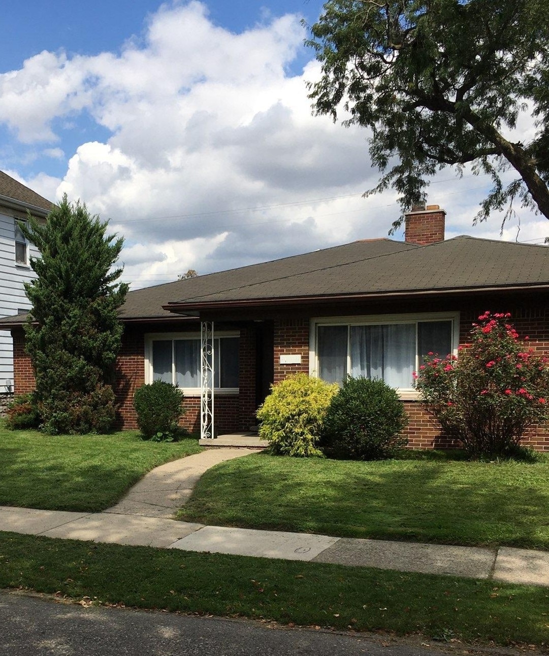 A single-story brick house is framed by a well-maintained lawn. The pathway leads to the entrance, flanked by lush shrubs and colorful flower beds. Large windows allow natural light to fill the interior, while a tall tree provides shade nearby.