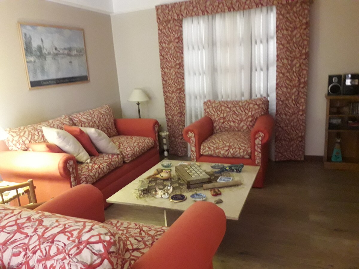 The living area showcases two red upholstered sofas with patterned cushions, arranged around a central coffee table adorned with decorative items. A small lamp stands beside a patterned curtain, creating a comfortable and inviting space. A wooden cabinet with audio equipment is visible in the corner.