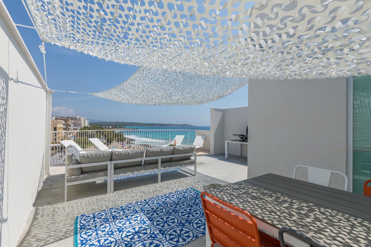 A spacious rooftop terrace is adorned with comfortable lounge chairs and a shaded dining area. A decorative outdoor rug adds a pop of color. The terrace provides views of the beach and the sea, offering a relaxing escape surrounded by a light canopy.