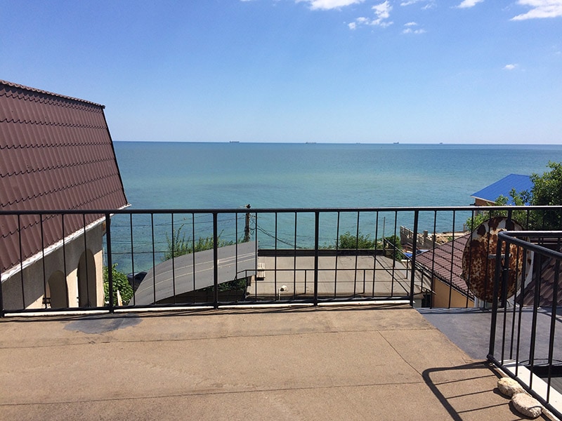 A terrace offers an unobstructed view of the Black Sea, with calm blue waters stretching to the horizon. The surrounding area features rooftops of nearby buildings, and sunlight reflects off the water, enhancing the serene coastal setting.