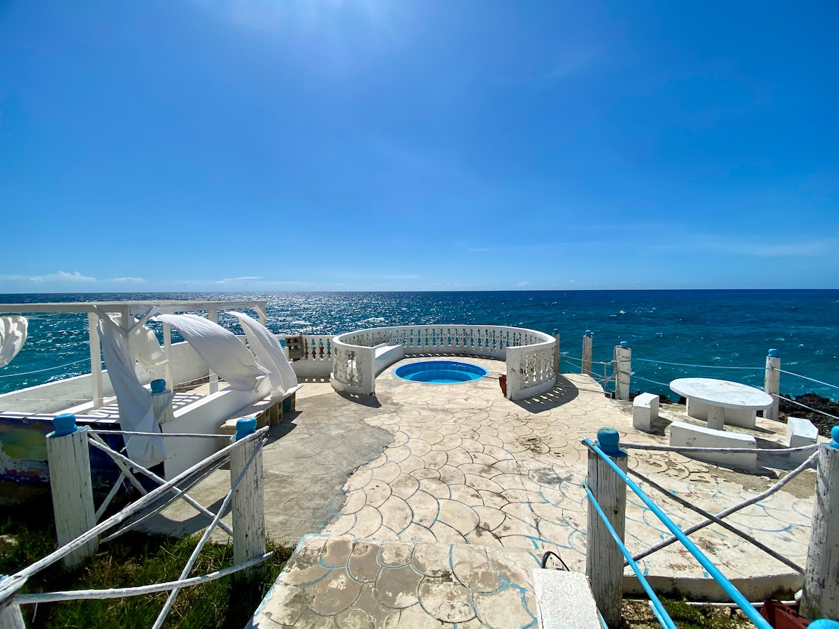 An outdoor terrace features a circular pool surrounded by white railing, offering panoramic ocean views. Several lounge chairs are arranged for relaxation, and a stone table sits nearby, creating a spacious area for gatherings. The bright blue sky contrasts with the shimmering sea.