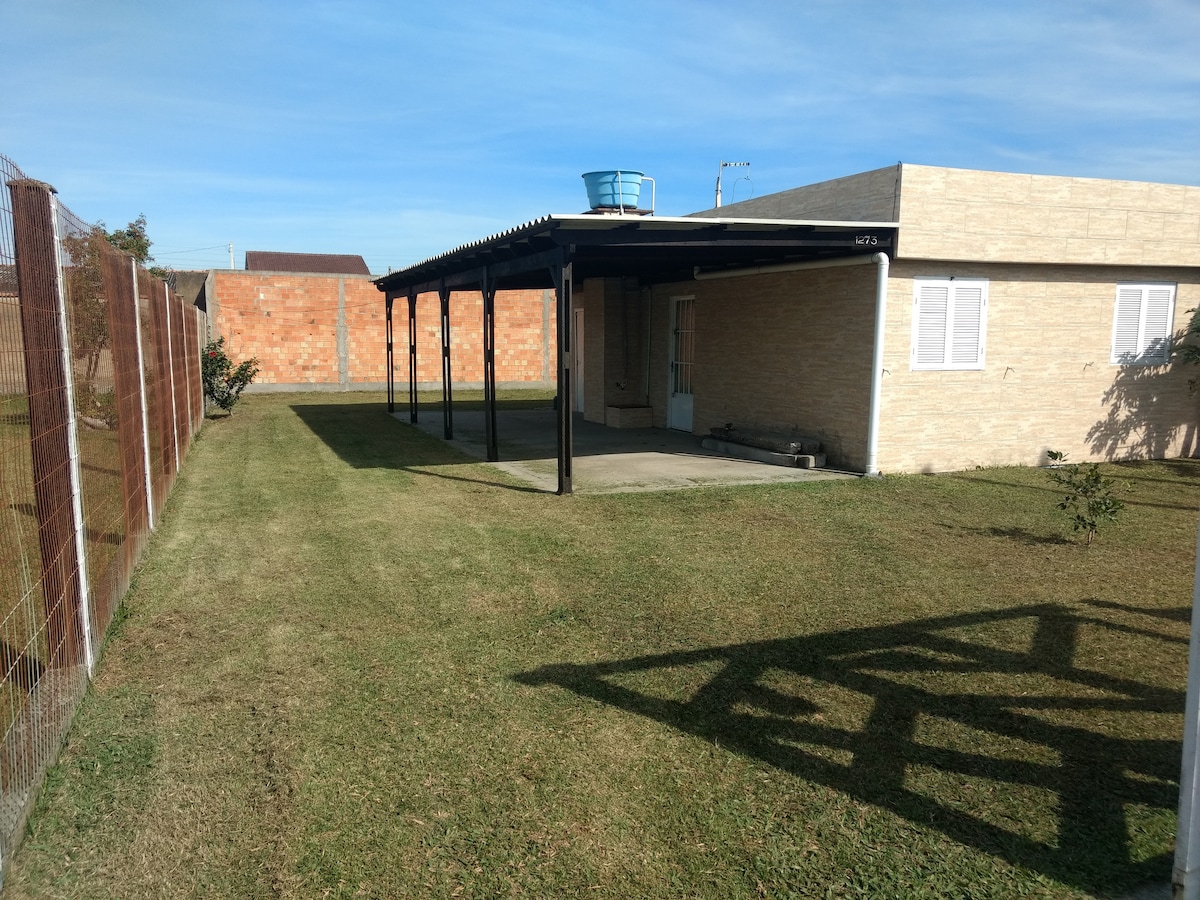 A modest house is set within a spacious green yard, bordered by a secure fence. A covered patio extends from the side of the house, providing shade. Behind, a brick wall and additional greenery can be seen against a clear blue sky.
