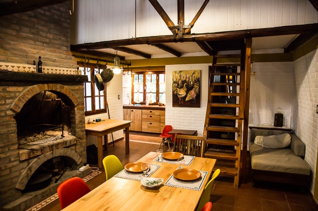 A rustic kitchen and dining area features a traditional brick oven and a large wooden dining table set with dishes. A spiral staircase leads to an upper level, while colorful chairs add a touch of vibrancy. Natural light enters through multiple windows, enhancing the space.