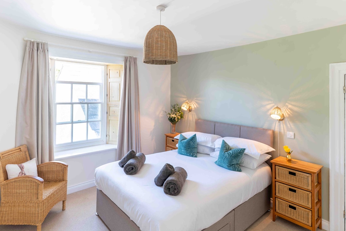 The tranquil bedroom features a spacious bed adorned with crisp linens and decorative pillows. Two light fixtures provide soft illumination, while a window framed by light curtains allows natural light to enter. A wicker chair stands nearby, accompanied by wooden nightstands offering storage.