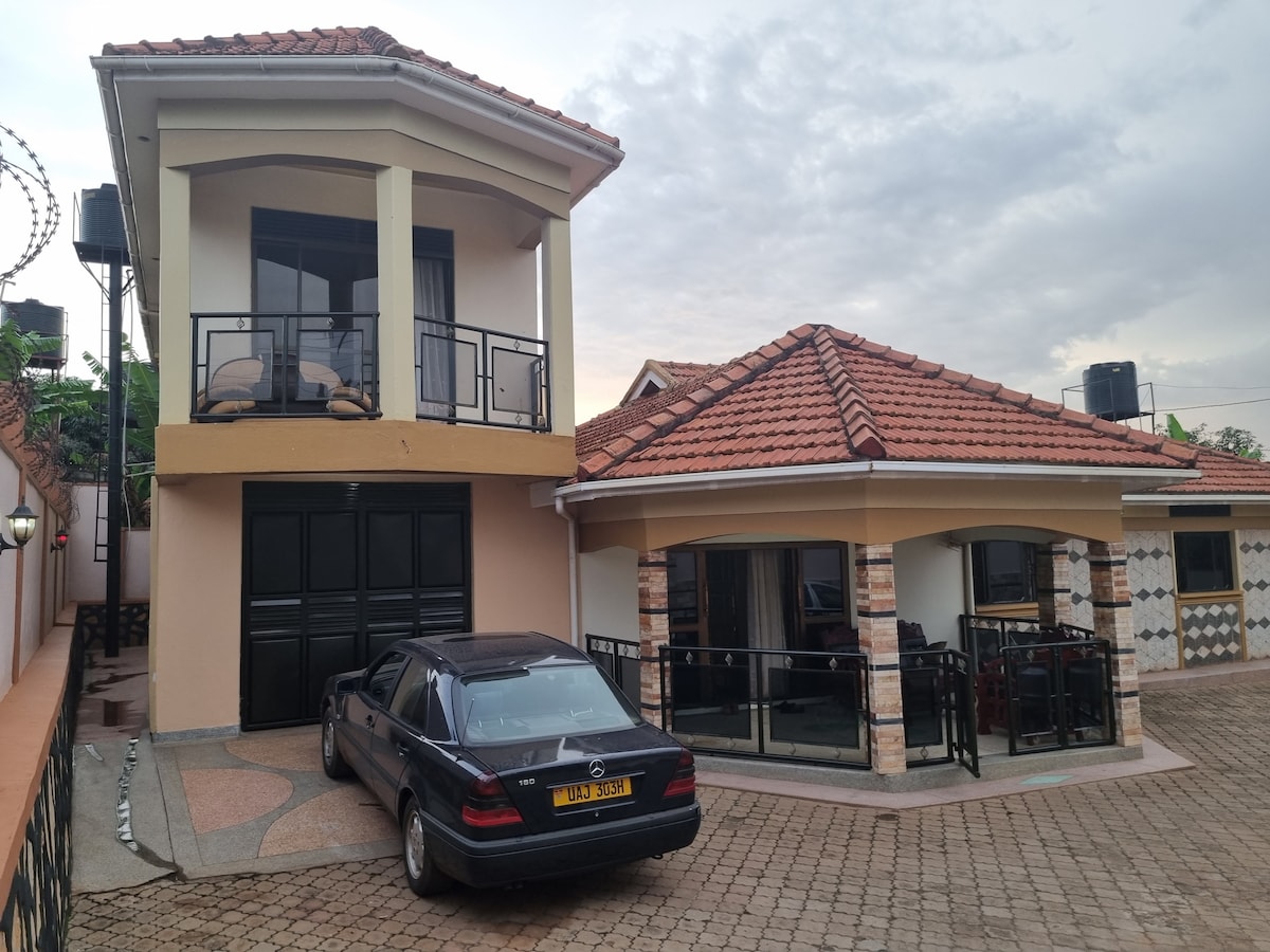 A two-story house is seen beside a single-story structure, both featuring terracotta tiled roofs. A parked black car is positioned in front, while balconies are visible on the upper floor. The exterior is complemented by stone accents and a paved courtyard.