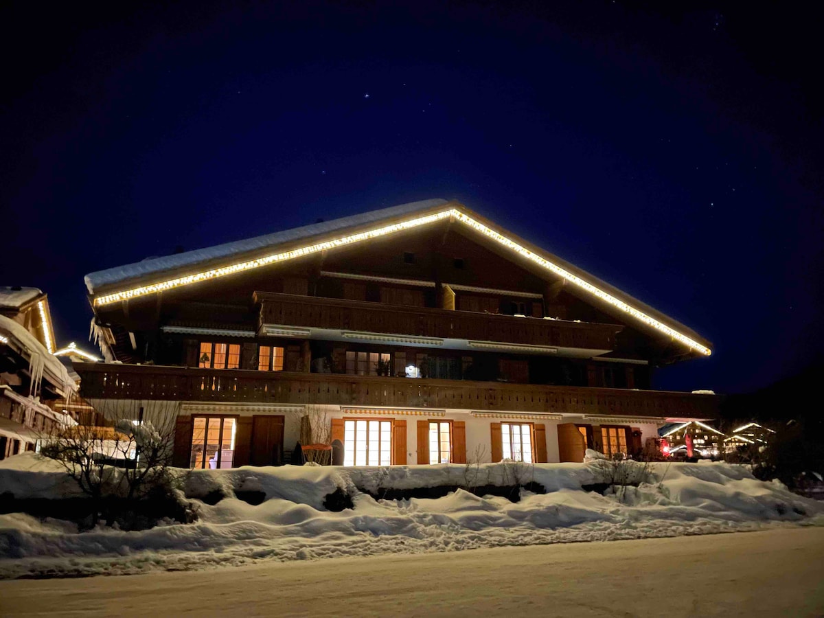 The chalet is illuminated at night, showcasing its wooden façade adorned with soft lighting. Snow blankets the ground, and warm lights from the windows suggest a welcoming interior. The structure features a spacious balcony and multiple levels, creating an inviting appearance against the night sky.