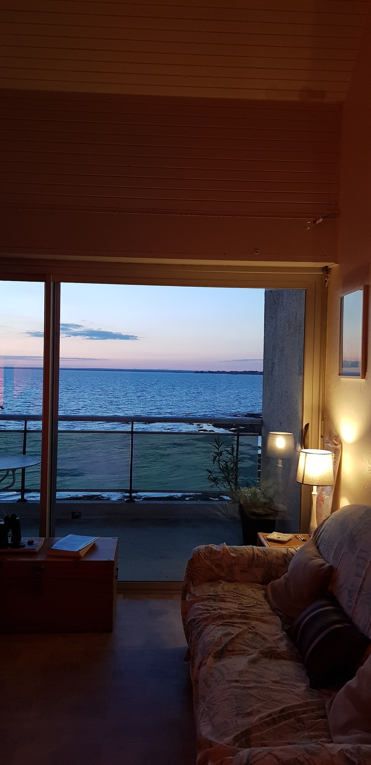 A peaceful living area is visible, showcasing a large glass door leading to a private balcony with ocean views. Soft lighting from a lamp offers a warm glow, while the horizon displays hues of blue and orange as the sun sets over the water.