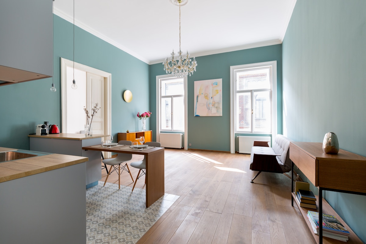 A modern living space features light wood flooring, complemented by a stylish dining table and chairs. Natural light enters through large windows, illuminating the blue-green walls. A decorative chandelier hangs from the ceiling, while a comfortable sofa and side table complete the welcoming atmosphere.