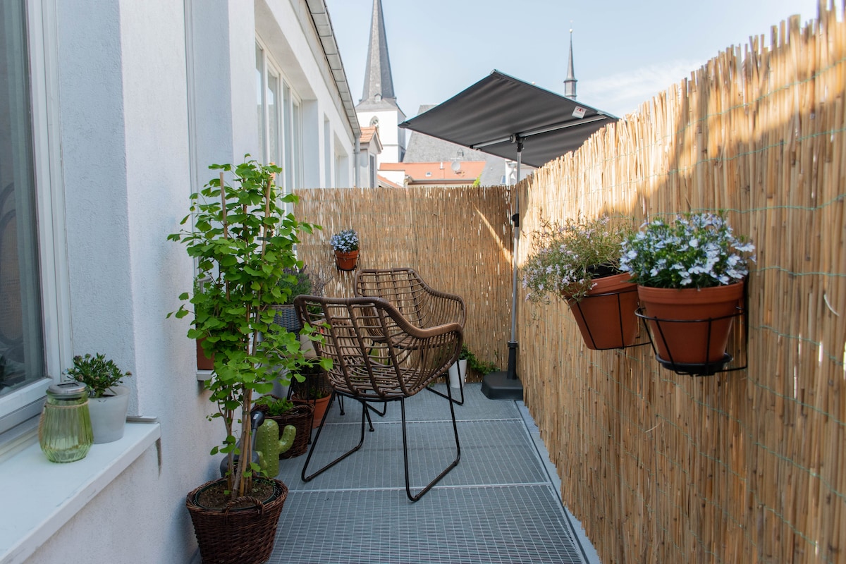 A private terrace offers a comfortable seating area with two woven chairs beside a potted plant. Hanging planters display blooming flowers, while a bamboo fence provides privacy. A sunshade is positioned overhead, and the surrounding cityscape features rooftops and church spires in the background.