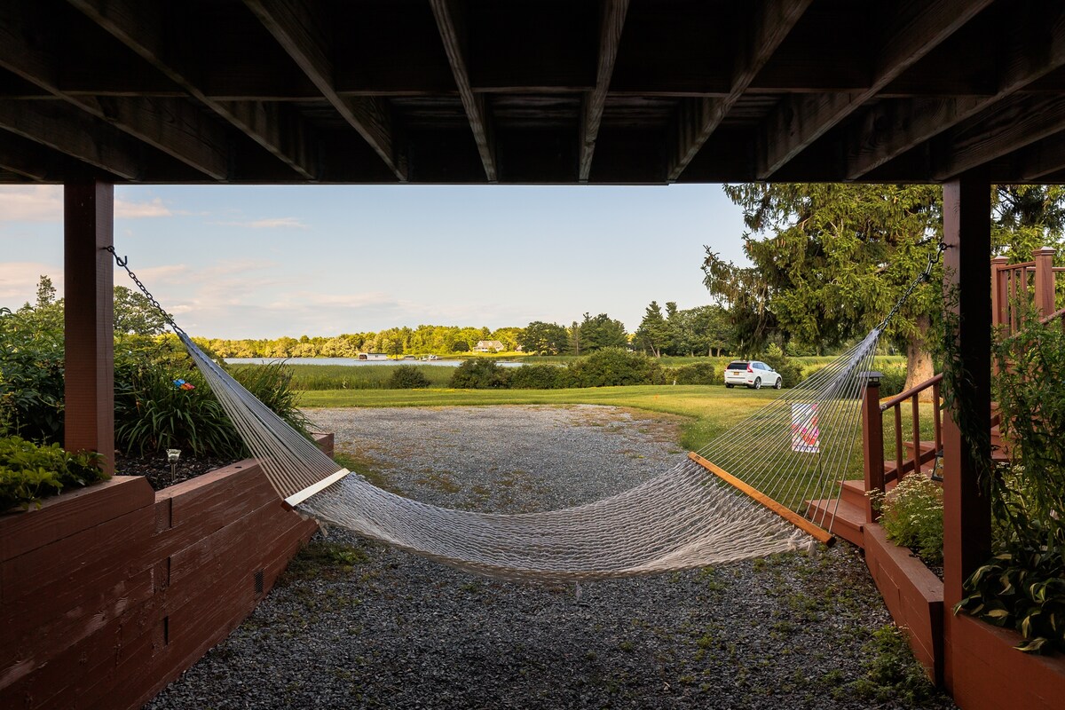 A hammock is suspended under the porch, facing a scenic view of greenery and the water beyond. The area is surrounded by gravel and landscaping, with a white car parked in a distant area. Natural light enhances the serene atmosphere.