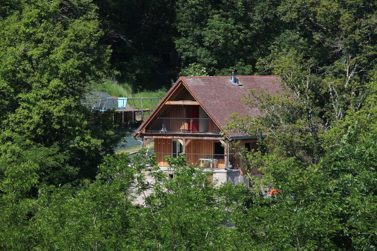 A wooden chalet is nestled among lush green trees, showcasing a sloped roof and large windows that allow natural light to enter. The structure is surrounded by foliage, creating a serene and private environment.