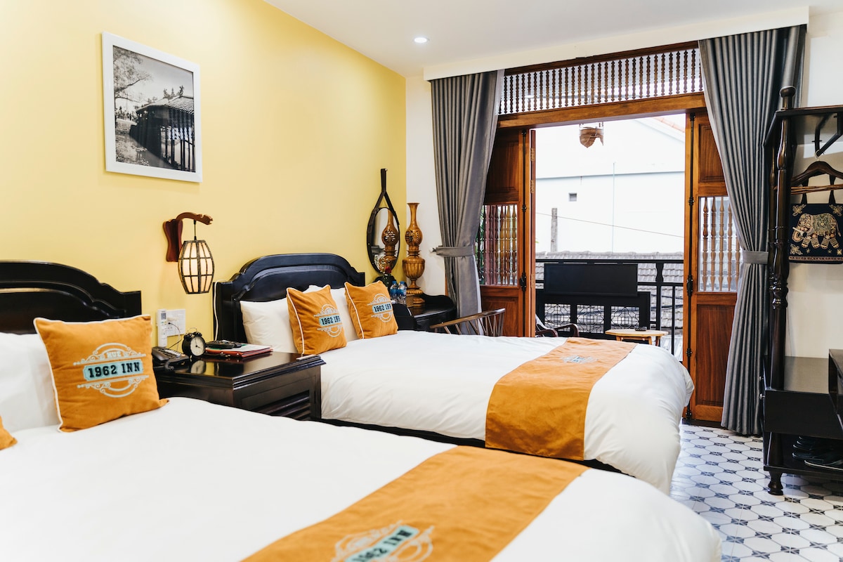 A well-appointed guest room features two spacious beds with orange and white linens. Decorative pillows are arranged on each bed. Natural light enters through large wooden doors that open to a small balcony, complementing the light yellow walls and patterned tiled floor.