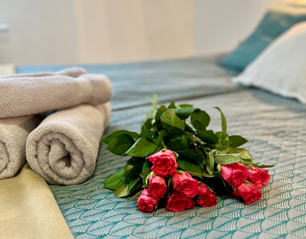 A neatly arranged bed features a soft, textured blanket and two decorative pillows. Rolled towels are placed beside a vibrant bouquet of pink roses, adding a touch of color to the inviting space.