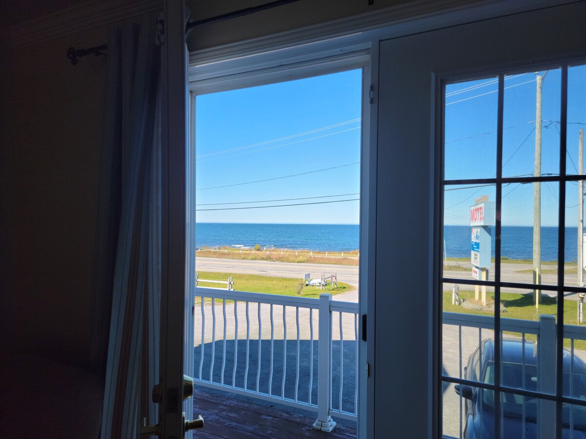 A view of the tranquil St. Lawrence River is visible through the open doors, showcasing a clear blue sky and gentle waves. A balcony railing frames the scene, while greenery and pathways are seen in the foreground, emphasizing the natural surroundings.