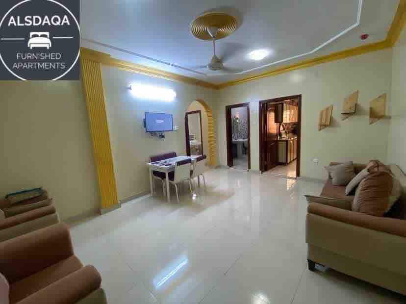 A bright living area features light-colored walls with yellow accents, complemented by a ceiling fan. A wall-mounted television is positioned above a small dining table, which is accompanied by two chairs. Comfortable seating is arranged on either side, with glossy tile flooring throughout.