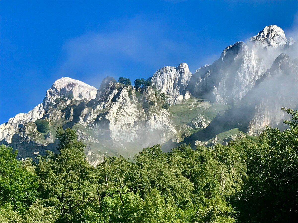 The rugged mountain landscape is characterized by steep rocky cliffs covered in patches of greenery. Wisps of mist are present, creating a serene atmosphere against the backdrop of a clear blue sky.