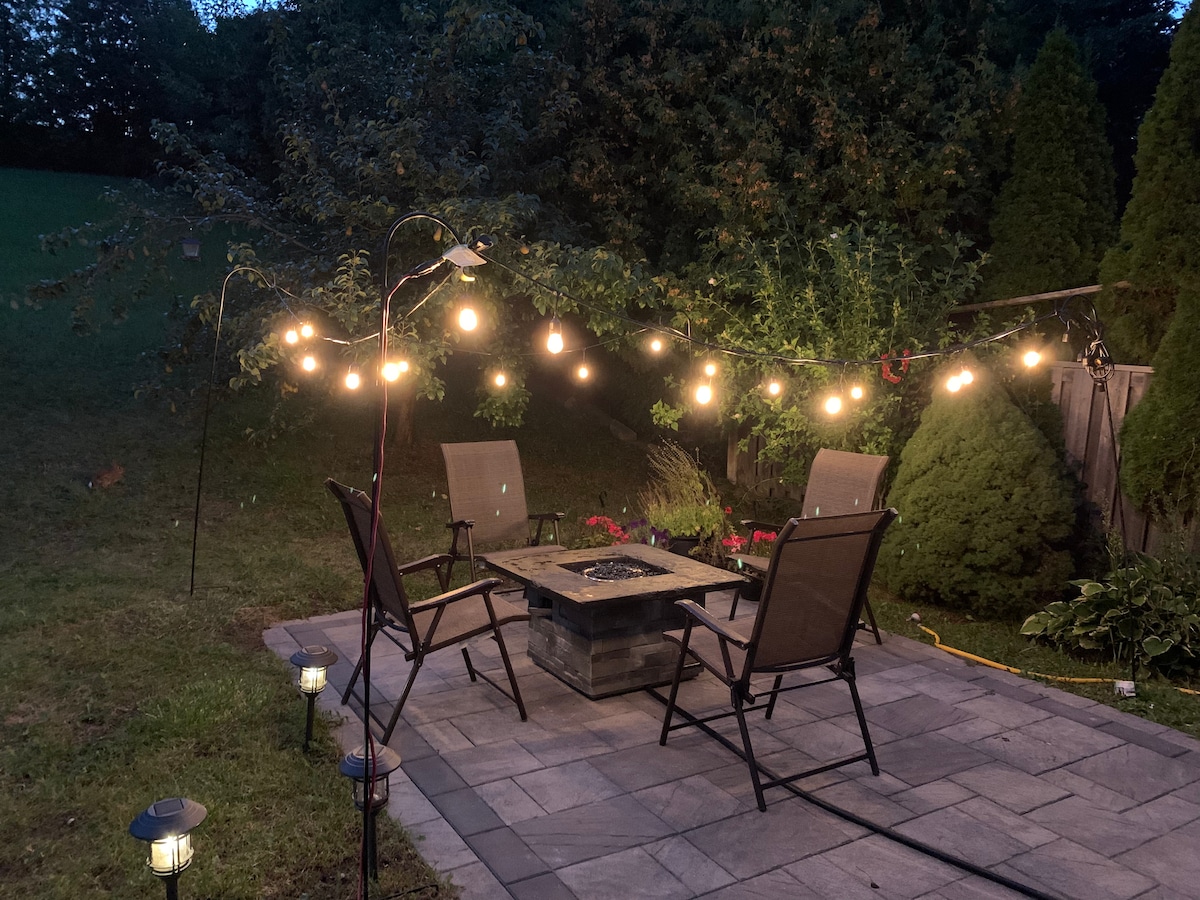 A cozy outdoor seating area is illuminated by string lights overhead. Four chairs are arranged around a square fire pit, surrounded by greenery and flower beds. Pathway lights provide gentle illumination, enhancing the inviting atmosphere of the space.