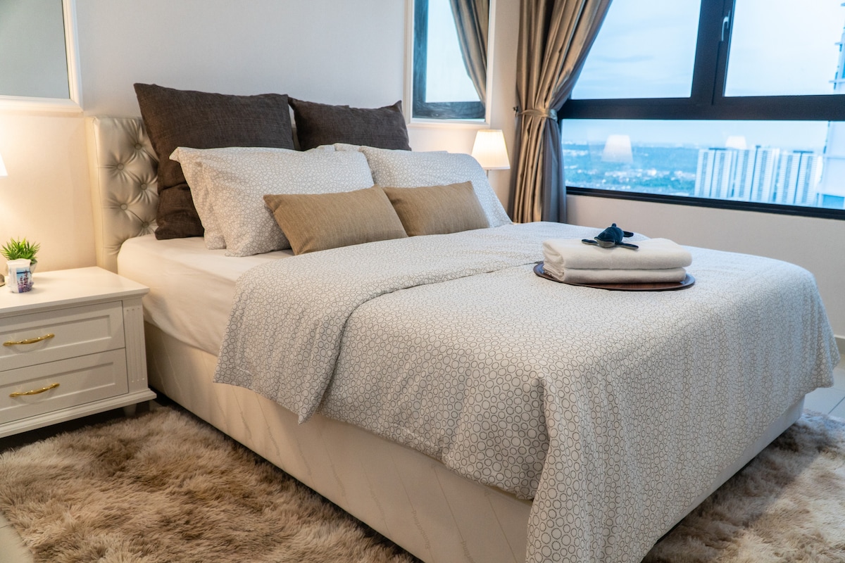 A spacious bed is dressed in soft, patterned linens with pillows in various shades. A small decorative plant sits atop a bedside table, and a plush area rug adds warmth to the room. Large windows provide a view of the cityscape beyond.