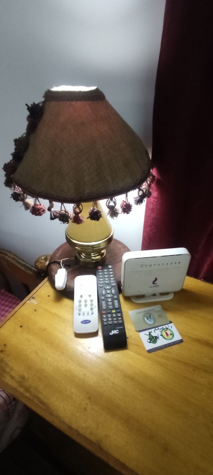 A wooden side table with a lamp featuring a shade adorned with decorative tassels. A remote control, a small electronic device, and various cards are neatly arranged on the table's surface, highlighting the functional details of the space.
