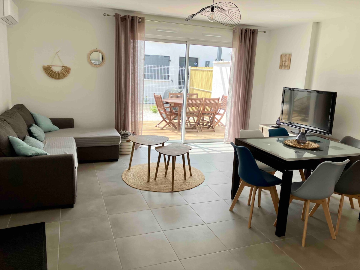 A bright living area is presented with a gray sofa and a pair of light blue accent cushions. A dining table with colorful chairs is positioned nearby, and large glass doors offer a view of a wooden terrace with outdoor dining furniture.