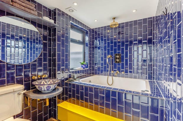 A modern bathroom is showcased with striking blue tiled walls and a large soaking tub. A stylish yellow bathtub adds a pop of color, complemented by a round mirror and a sink with decorative detailing. Natural light filters in through a small window.