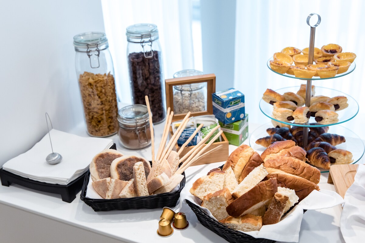A breakfast spread is arranged on a white surface, featuring assorted baked goods, including various types of bread and pastries. Clear glass jars contain cereals and dried fruits, while small cartons of drinkable items are visible. Wooden serving utensils and napkins are also present.