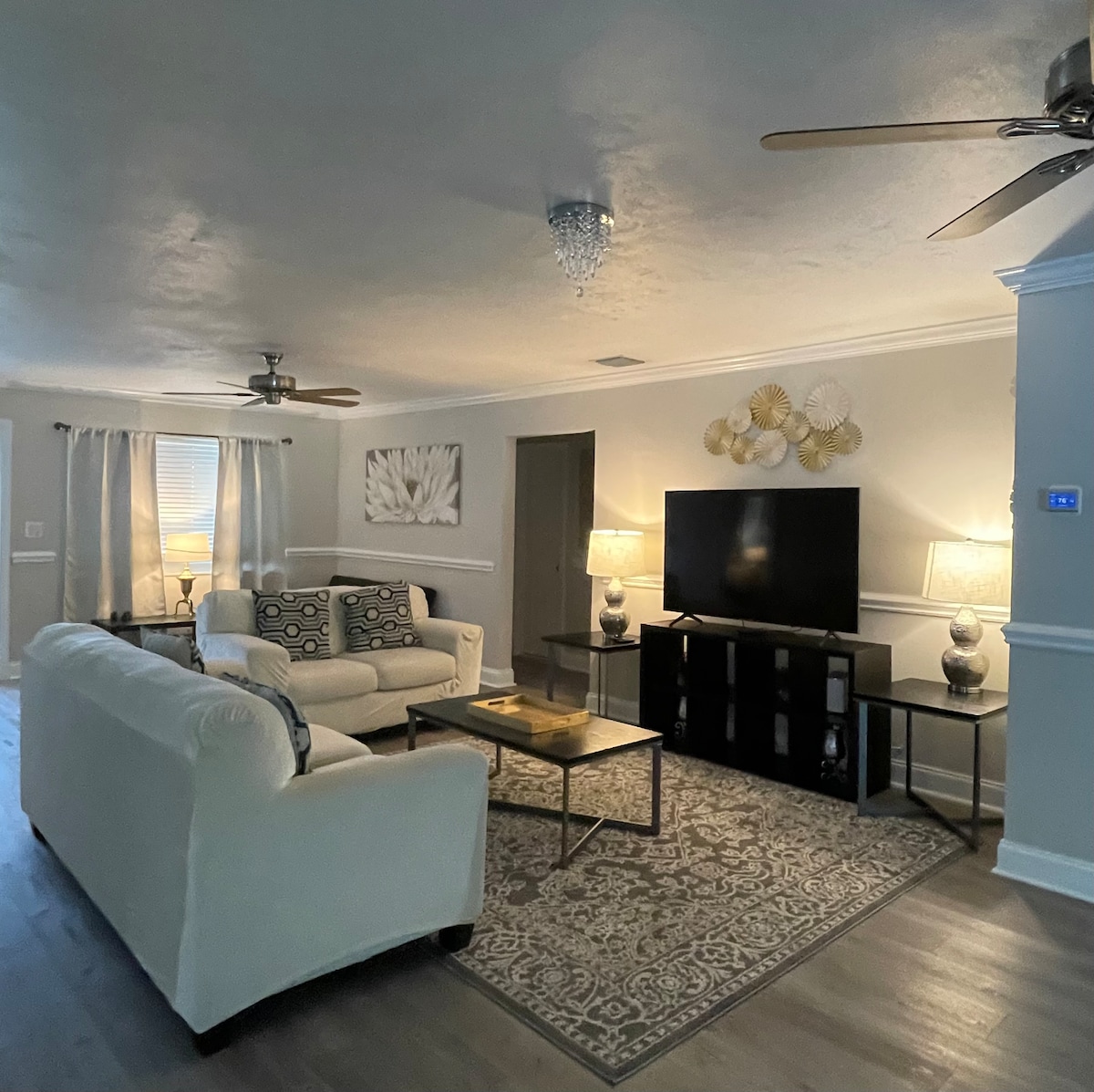 The living room showcases a neutral palette with two light-colored couches and a coffee table in the center. A large smart TV is positioned on a dark media console against the wall, complemented by soft lighting from two lamps and ceiling fans above.