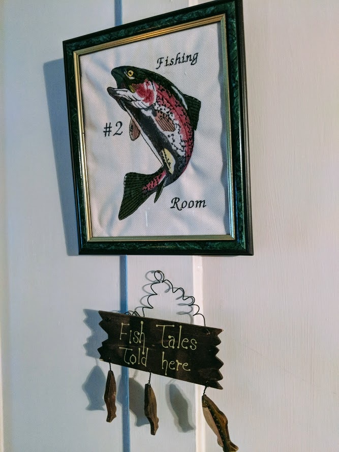 A framed artwork featuring a fish is displayed on the wall, labeled 'Fishing Room #2'. Below, a rustic sign hangs, stating 'Fish Tales Told Here.' The arrangement adds a thematic touch to the decor in the lodge.