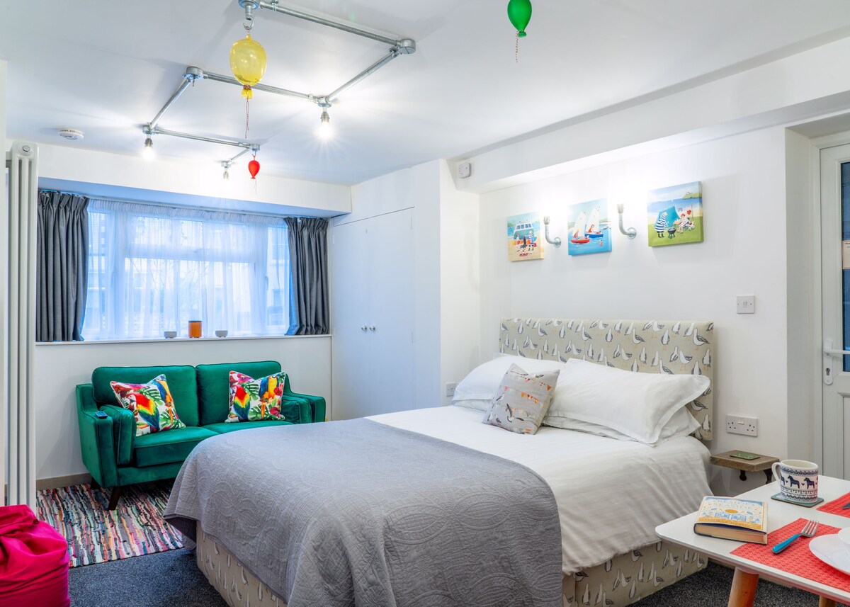 A cozy studio features a spacious bed dressed in neutral bedding. A bright green cuddler sofa with colorful cushions sits beside a window, providing natural light. Cheerful wall art adds a touch of color, and decorative lighting fixtures create a warm ambiance.