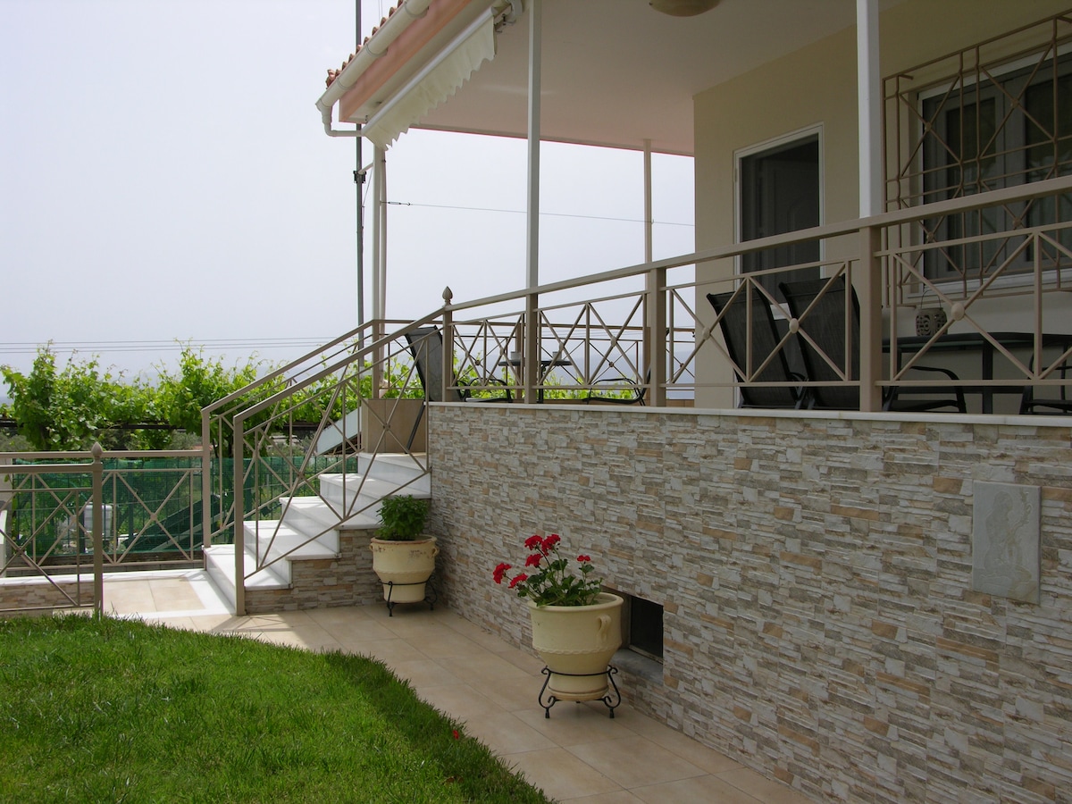 A well-maintained entrance features a gentle sloped ramp leading to a spacious covered terrace. The façade showcases a mix of stone cladding and light-colored tiles. Potted flowers add a touch of color, while a railing frames the space, ensuring accessibility and aesthetic balance.