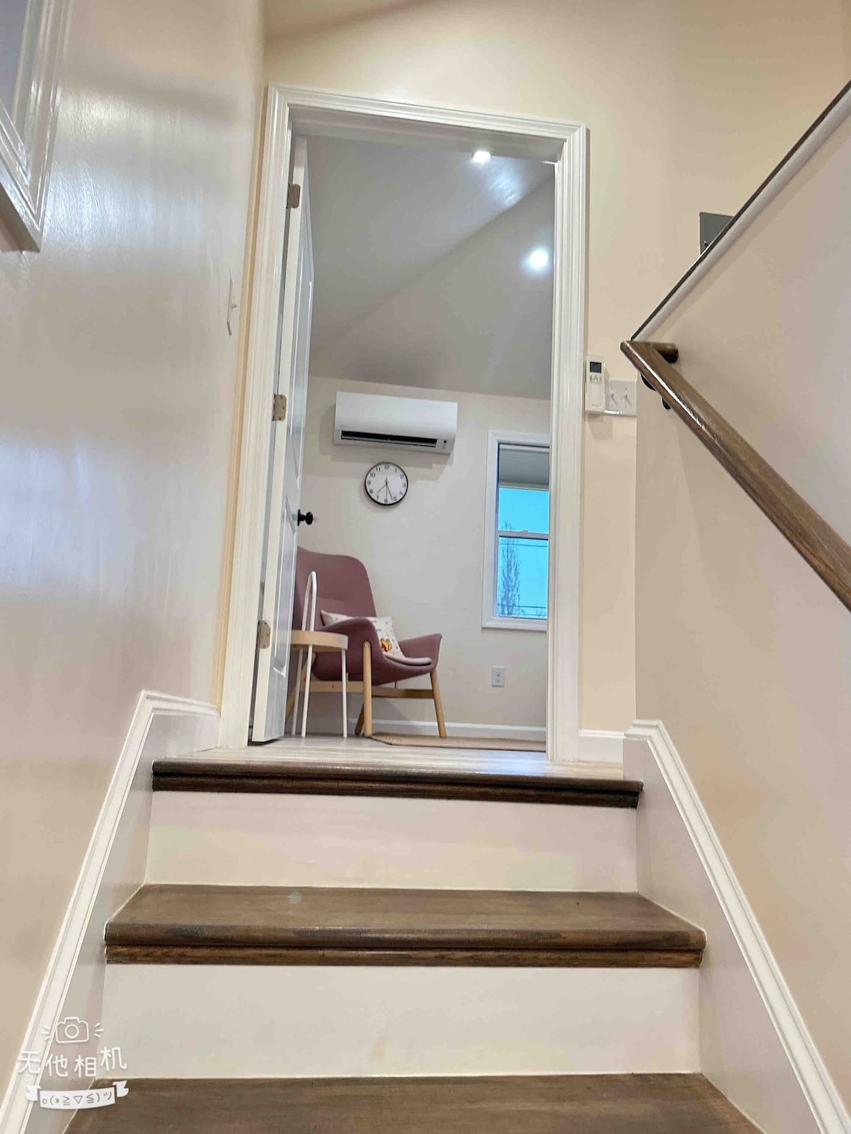 A staircase leads to an upper level, featuring light wooden steps and white walls. A door with a glass pane opens into a room that includes a clock on the wall and a chair positioned near a window, allowing natural light to enter.