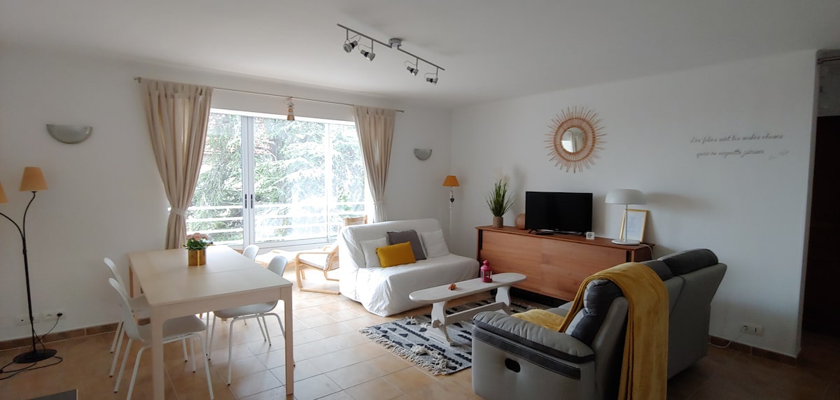 The spacious living and dining area features large windows with natural light and views of greenery. A comfortable sofa and armchair, alongside a minimalist dining table, create a welcoming ambiance. A wooden media unit houses a TV, while decor accents add subtle charm.
