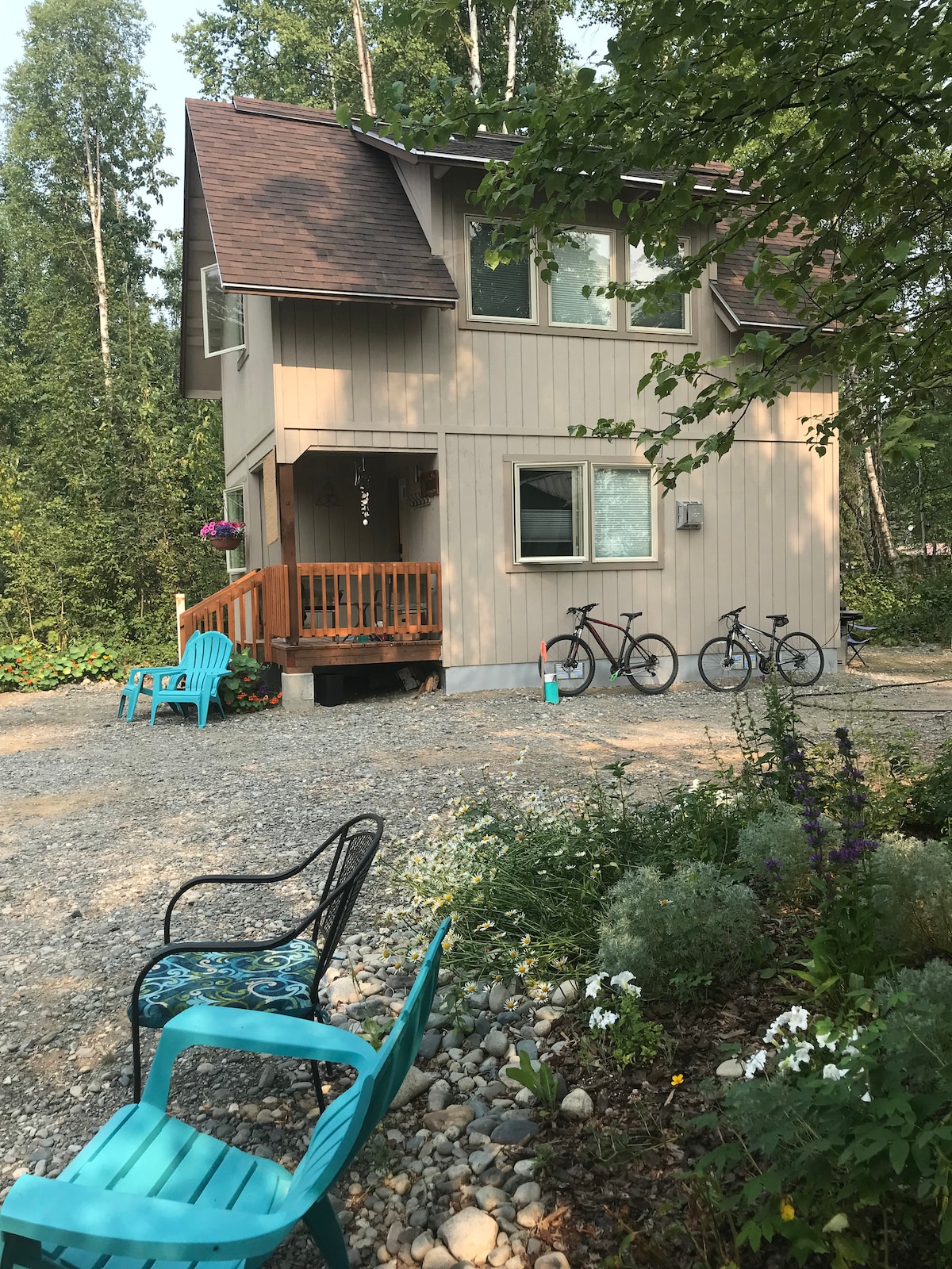 A two-story house is set amidst a natural landscape, featuring a wooden porch and vibrant hanging plants. Two bicycles are parked nearby, and a gravel path leads to seating areas with bright chairs. Lush greenery and flowers surround the property, creating a refreshing outdoor space.