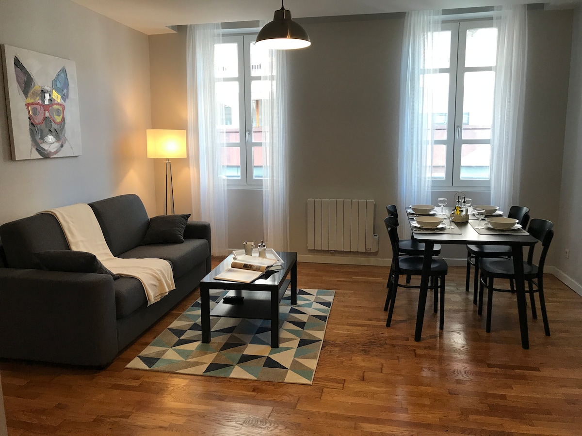 A spacious living area features a comfortable gray sofa adorned with a light throw. Light streams in through large windows draped in sheer curtains. A dining table is set for four, situated on a stylish area rug that complements the wooden floor.