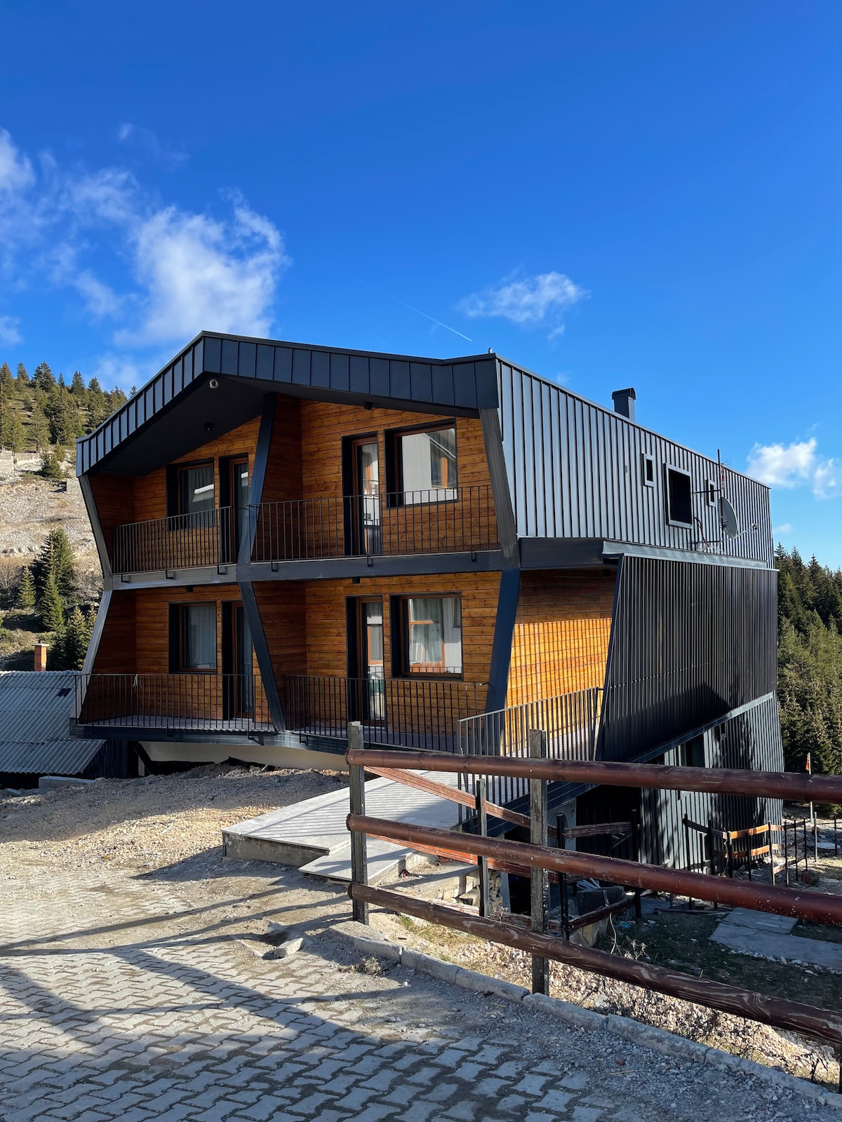 A multi-level, modern building is surrounded by a natural landscape, featuring a combination of wooden and metallic exterior finishes. Balconies adorn each level, offering a connection to the outdoors. Clear skies and scattered clouds provide a serene atmosphere.