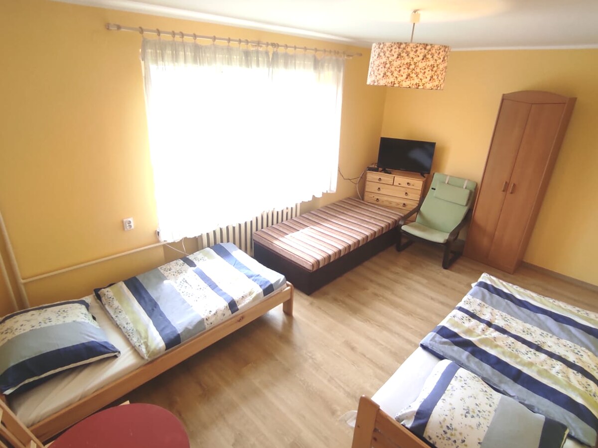 A guest room is presented with three beds featuring patterned bedding. A television is mounted on a wooden dresser beside a long, cushioned bench. A comfortable armchair is positioned near a large window, allowing natural light to fill the space. The walls are painted in a soft yellow.