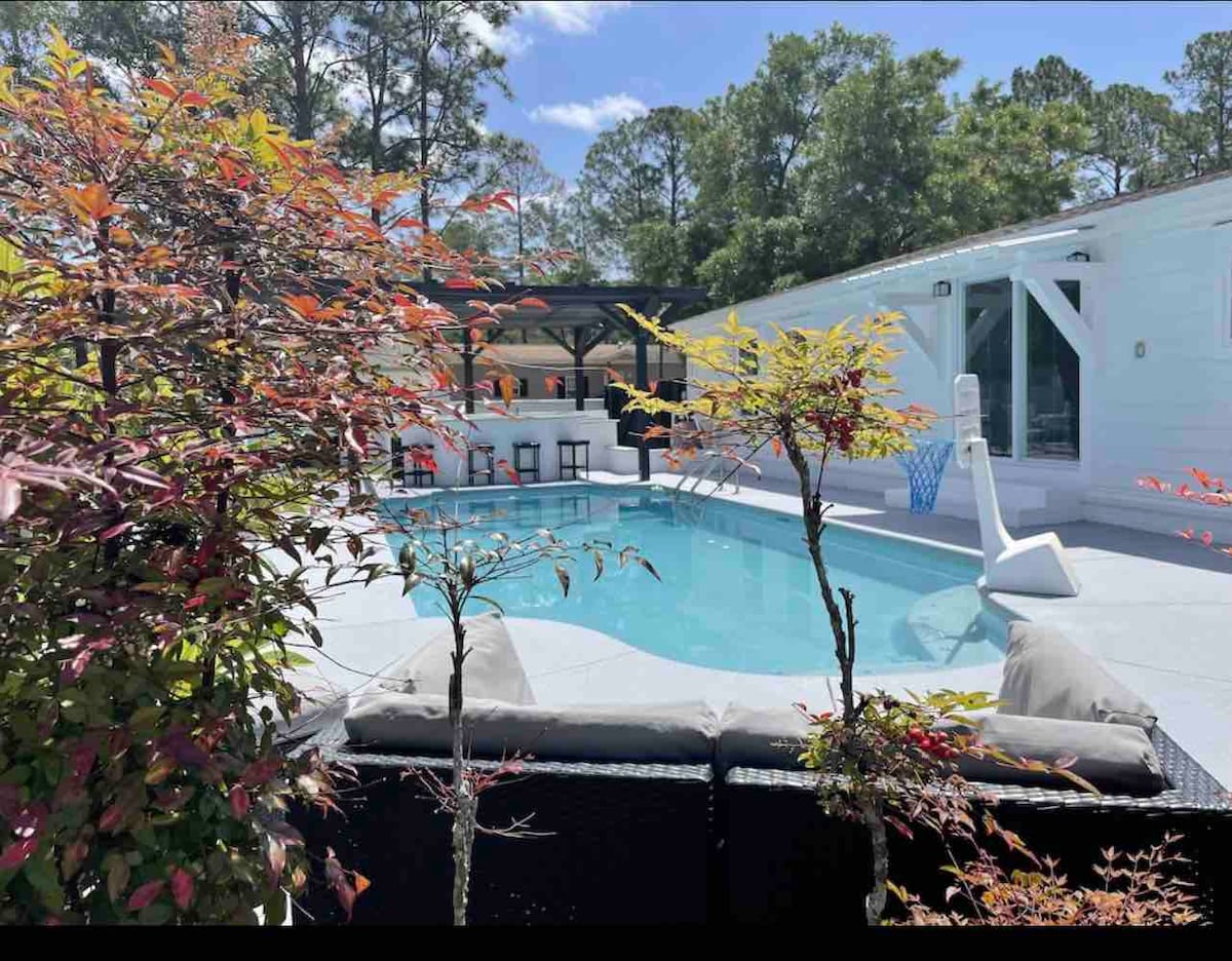 A private swimming pool is surrounded by lush greenery and vibrant foliage. Lounge seating is visible nearby, and a covered outdoor bar area provides a shaded space for gatherings. The pool area is brightly lit by natural sunlight, creating a welcoming outdoor retreat.
