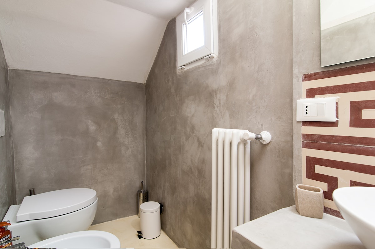 A modern bathroom is presented with a minimalist design. Textured gray walls complement a compact toilet and washbasin. A small window provides natural light, while a towel radiator and a waste bin are included. Simple decor elements enhance the functional space.