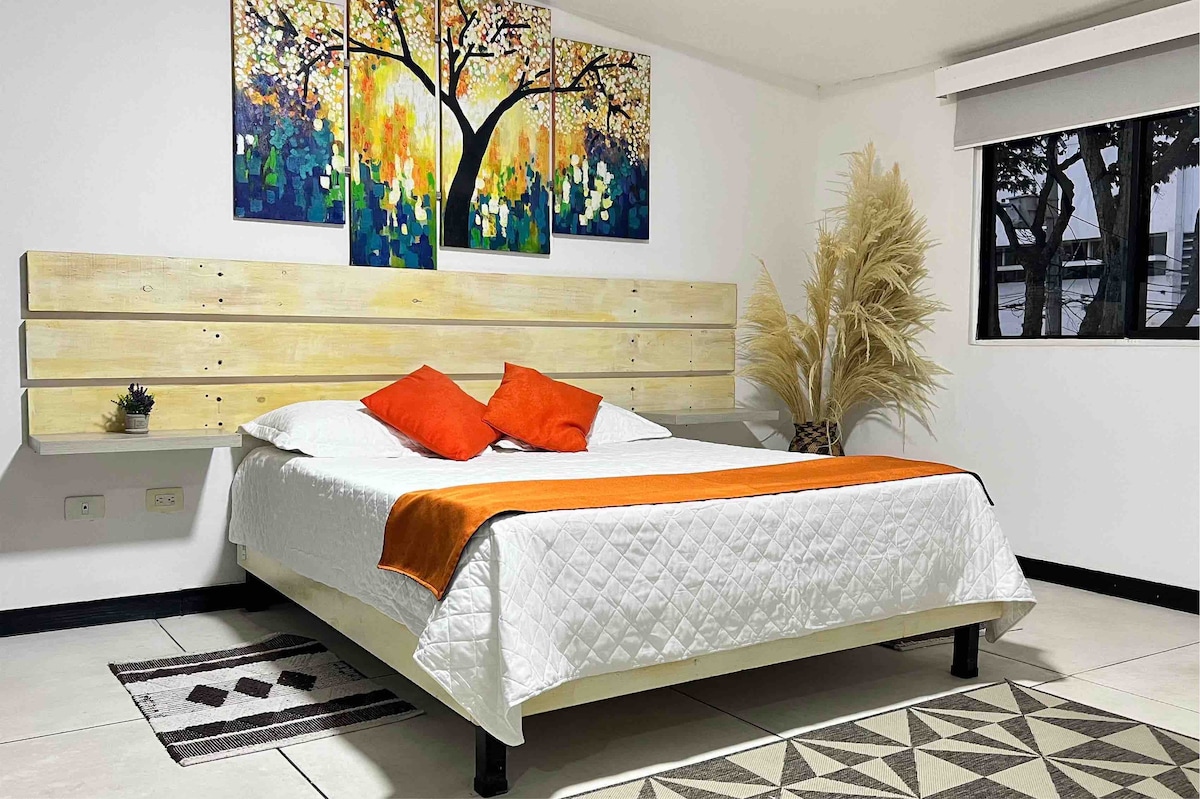 A spacious sleeping area features a Queen bed with a white quilt and bright orange accents. The headboard is made of wooden planks, while a vibrant triptych of artwork is displayed above. A decorative plant stands nearby, and natural light filters through a window.